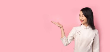 Geleneksel elbiseli mutlu gülümseyen Çinli kadını pembe arka planda sol tarafa bakarken gösteriyor. Kameraya bakar ve gülümser. uzun yatay afiş kopyalama alanı