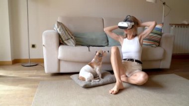 Sarışın kadın VR kulaklık takıyor. Oturma odasında, yerde, küçük köpeği Jack Russell Terrier ile oturuyor. Köpek, sahibini kafa karışıklığı ve bulmacayla izliyor. Sanal gerçeklik video görüntüleri