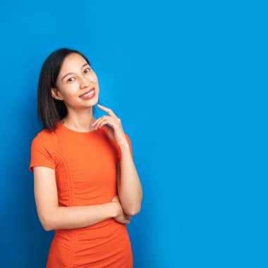 Chinese woman in orange dress looking in camera and smiling. Blue background. Banner. thoughtful pose, hands on the waist, surprise. square composition