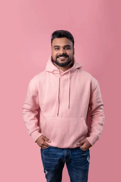 Relaxed Men in pink clothes. Smiling handsome Pakistani guy looking at camera. Casual pink hoodie hands in pockets. Attractive smile. Pink background. Positive mood. Template clothes
