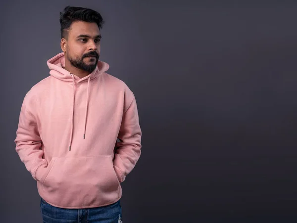 Man in pink hoodie on grey background. Handsome modern young Pakistani guy looking side. Casual pink hoodie hands in pockets. Yellow background. Serious pensive face expression. Gray empty copy space