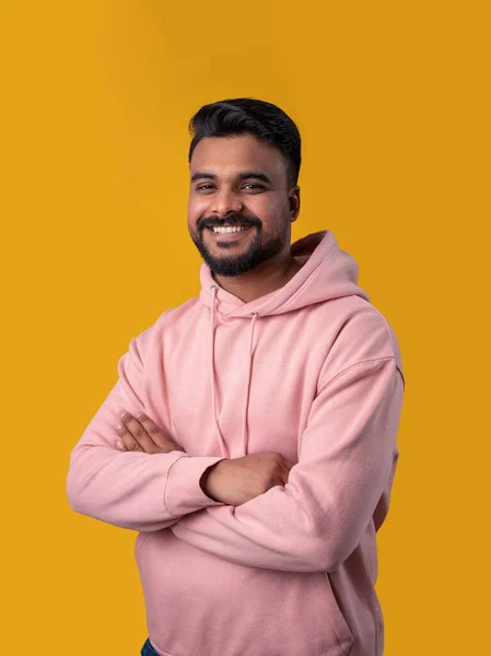 Smiling handsome Pakistani guy looking at camera. Casual pink hoodie crossed arms. Attractive toothy smile. Yellow background. Positive mood