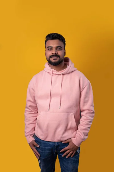 Men in pink clothes. Smiling handsome Pakistani guy looking at camera. Casual pink hoodie hands in pockets. Attractive smile. Yellow background. Positive mood. Template clothes