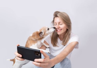 Kadın ve köpek selfie çekiyor. Küçük köpek Jack Russell Terrier cep telefonuyla sarışını öpüyor. Beyaz elbiseler, gri, sağlam arka plan. Sosyal medya içerik oluşturucu