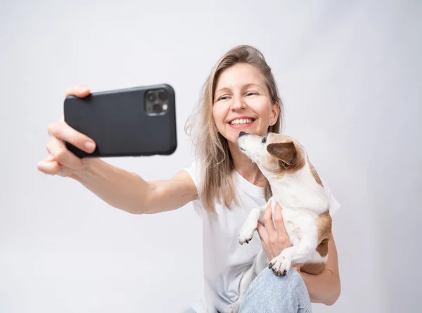 自私自利的金发女人和她可爱的小狗小狗杰克罗素亲吻金发碧眼的女人手持手机 白色衣服背景灰坚实 社交媒体内容创建者 — 图库照片