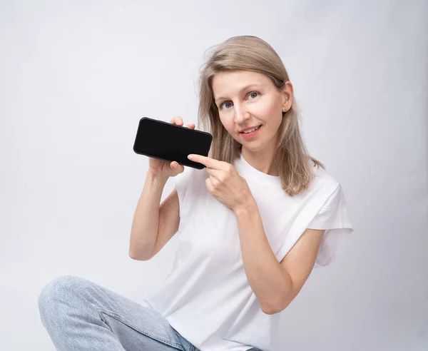 Atraktivní Blondýnka Ukazující Černý Mobil Ukazující Aplikaci Bílé Tričko Modré — Stock fotografie