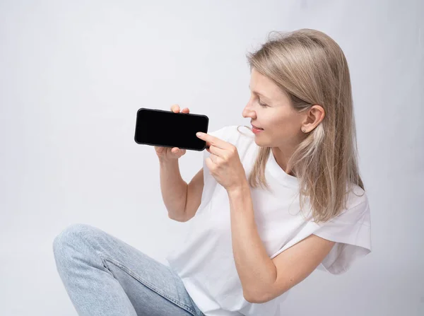 Woman side profile pointing finger on black cell phone showing the application. White t-shirt blue background. Happy relaxed calm smile. Gray background one person. Holding using cell phone