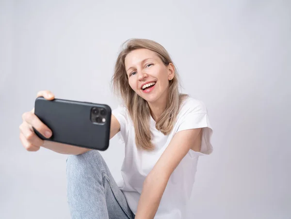 Blonde woman holding phone taking selfie chatting online video call. White t-shirt blue background. Happy wide smile. Gray background one person. Holding using cell phone