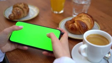Kromakey yeşil ekran eli yatay olarak cep telefonunu tutuyor. Oyunu oynarken el sıkışması. Kahveli ve kruvasanlı sokak kahvesi. Şablon video görüntüleri 