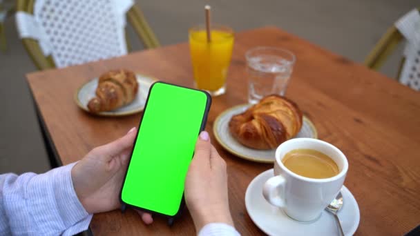 携帯電話の縦位置を保持するクロマキーグリーンの画面の手 指でソーシャルメディアをスクロールします ダブルクリックします テンプレートビデオ映像 コーヒーとクロワッサンと一緒にストリートカフェで朝食 — ストック動画