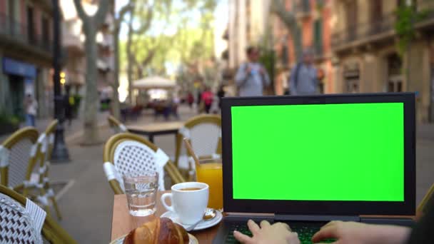 ストリートカフェで働く緑のクロマキー画面とノートパソコン リモートワークやオンラインショッピング 背景を模倣しろ クロワッサンとテーブルの外 コーヒーと水のカップ ビデオ映像 — ストック動画