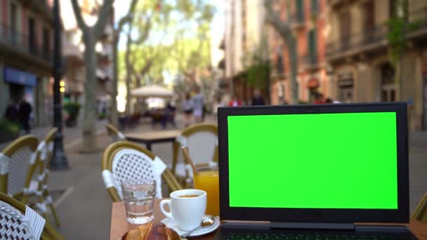 ヨーロッパのストリートカフェのテーブルの上に緑の画面を持つノートパソコン 手をタイプするな 静的シーン クロマキーモックアップ付きのテンプレートビデオフッテージノートブック バルセロナでクロワッサンとモーニングコーヒー — ストック動画