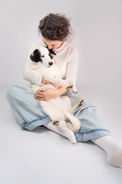 Dog owner and dog cuddling. Hugging young woman and adorable white border collie pet sitting on the floor. grey (gray) background. Casual blue jeans and white clothes. Best friends trust relationship