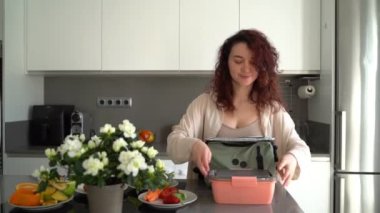 Kıvırcık saçlı genç kadın pembe beslenme çantasını mavi çantaya koyup çalışmaya ya da işe gitmeye hazırlanıyor. Meyve ve çiçekli güneşli modern mutfak. Her gün sağlıklı alışkanlıklar video görüntüleri.
