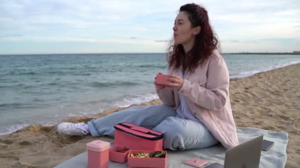 海の近くで昼休み ビーチで昼食を取った若い女性がプラスチックピンクの食品容器を持って食べています 風の強い昼休み ビデオ映像を食べる — ストック動画