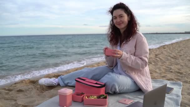 ビーチでランチをした若い女性は プラスチック製のピンクの食品容器からチョコレートクッキーを食べています 風の強い昼休み ビデオ映像を食べる — ストック動画