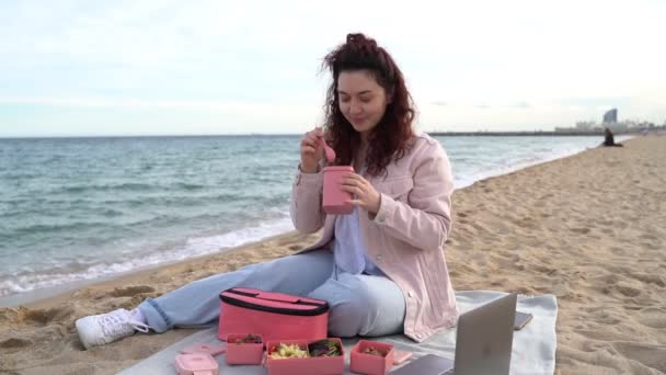 在海滩吃午饭的年轻女人吃着塑料粉红食品容器里的汤 刮风的午休 在外面吃饭的录像 — 图库视频影像