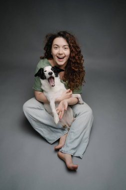 Sınırdaki köpeğini kucaklayan mutlu kadın gülerek kameraya bakıyor. Gri arka plan stüdyo. Yerde mavi kot pantolonla çıplak ayakla oturmak.