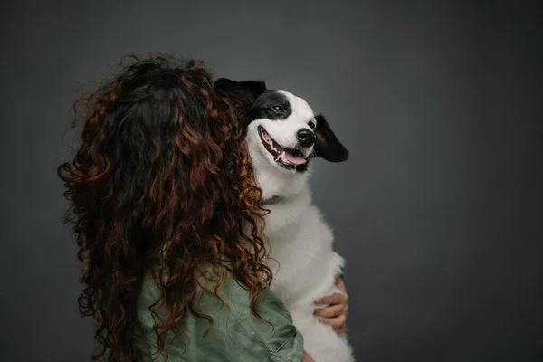 Back of woman with long curly hair holding her white border collie dog happy smiling and looking at the camera. Grey background studio shot.