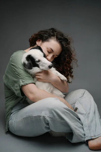 anxious dog hugs.  Beautiful woman with curly hair calming hugging cute sad dog sitting on the floor with dark grey background. Pet mental support. Blue jeans barefoot .