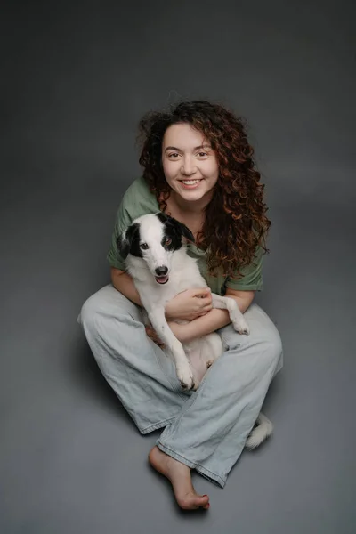 ハッピーな女性は彼女の国境のコリー犬を抱きしめ 笑ってカメラを見ています 愛する友人の肖像画 グレイバックスタジオショット ブルージーンズの床に座って裸足 — ストック写真
