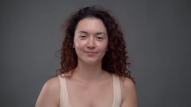 30 yaşında güzel bir kadın. Parlak tenli ve kıvırcık saçlı. Kameraya bakıyor ve gülümsüyor. Gri arka plan. Güzellik Sağlığı Pozitif temalı video görüntüleri