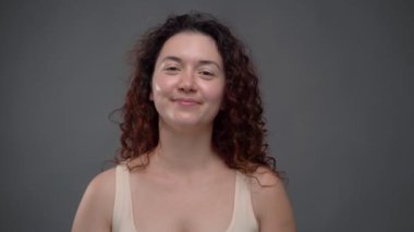 30 yaşında güzel bir kadın. Parlak tenli ve kıvırcık saçlı. Kameraya bakıyor ve gülümsüyor. Gri arka plan. Güzellik Sağlığı Pozitif temalı video görüntüleri