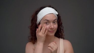 Rutin cilt bakımı. Kendine zaman ayır. Kafasında beyaz bandaj olan kadın cildine krem sürüyor. Gri düz arka plan stüdyo çekimi. Uzun kıvırcık saçlı. Keyfim yerinde