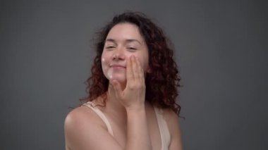 30 yaşında güzel bir kadın yüzüne kozmetik serumu sürüyor. Cilt bakımı yüz masajı rutin video görüntüleri. Gri arka plan stüdyosundaki kıvırcık saçlı beyaz kadın. Gülümseyen olumlu duygular