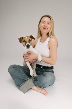 Gülen sarışın kadın ve köpek kameraya bakıyor. Stüdyo portresi. Köpek sahibi aşkı. Kameraya bakıyorum. Gri arka plan. Mavi kot beyaz gömlek