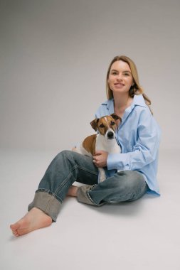 Mavi gömlekli, gülümseyen sarışın kadın yerde oturuyor, köpek kucağında oturuyor. Komik şirin arkadaşlar stüdyo resmi. Gri arka plan. Dikey bileşim