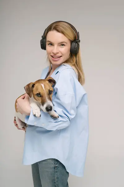 ハッピーミュージック愛好家ブロンドの若い女性は カメラを見ている小さな犬ジャックラッセルテリアを保持しています スタジオショット グレーバック ロイヤリティフリーのストック写真