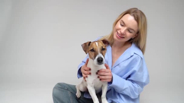小さな犬ジャックラッセルテリアを持つかなり若い金髪の女性の所有者 カメラを見て ペットを連れて笑顔になる スタジオビデオ映像 ハッピーフレンド — ストック動画
