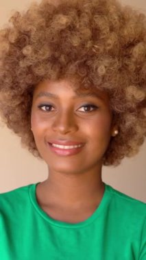 Afrika tarzı saçı olan, yeşil tişörtlü, kameraya bakıp gülümseyen mutlu siyah bir kadın. dikey video görüntüleri. Zarif hafif makyaj, inci küpeler.