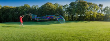 Yeşil yaz arka planında sabun köpükleri. Gökkuşağı renginde sabun köpükleriyle eğlenen kırmızı elbiseli kadın. Tatil havası için boş fotokopi alanı olan uzun yatay yaz anları afişi
