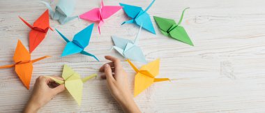 Origami sanatı. Arka planda beyaz ahşap masa bulunan kağıttan renkli origami vinçleri tutan eller. Katlama ve rahatlatma teknikleri yoluyla kağıt heykel