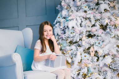 6 yaşında, güzel, küçük bir kız. Mavi sandalyede modern beyaz Noel ağacının yanında oturuyor. Şimdiki kutusunu açmak için bekliyor. Mutlu anlar kış aile tatilleri
