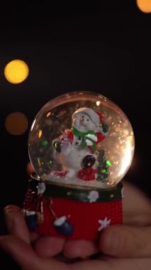 Kış tatili dekorasyonu kristal kar topu içinde kadın elinde komik bir kardan adam. Koyu siyah arka plan ve Noel ışıkları. Yavaş çekim dikey video görüntüleri. 