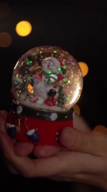 Ellerinde kristal kardan adam tutan bir kadın. İçinde komik kardan adam var. Koyu siyah arka plan ve Noel ışıkları. Yavaş çekim dikey video görüntüleri. 