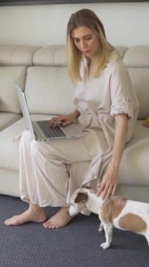 Sarışın kadın oturma odasında kanepede oturuyor kucağında dizüstü bilgisayar ve yanında köpek oturuyor. Evcil bir arkadaşı okşamak. Dikey video görüntüleri