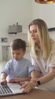 Sarışın kadın ve küçük çocuk kanepede dizüstü bilgisayar kullanarak oturuyor. Çocuk cihazları kullanmayı öğreniyor. Dikey video görüntüleri. Çocuk yetiştirmek.