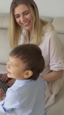 Genç bir anne dizüstü bilgisayarla kanepede oturuyor ve 3 yaşında bir çocuk yaklaşıyor. Kameraya bakarak mutlu bir şekilde gülümsüyor. Aile ortamı. çocuk yetiştirmek için evden çalışmak