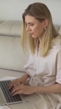 Dizüstü bilgisayarlı, bej koltukta oturan güzel sarışın kadın küçük köpeğe Jack Russell Terrier diyor. Köpek yaklaşımı ve ev hayvanı. Dikey video görüntüleri. Evden çalışmak rahat bir ruh hali. 