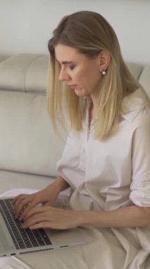 Kanepede oturan sarışın kadın dizüstü bilgisayarla çalışıyor ve yanında küçük köpek Jack Russell Terrier oturuyor. Dikey video görüntüleri. Tedbirli, şık bej renkler.