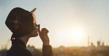 Siyah şapkalı genç kız. Gün batımında Avrupa şehri panoramasında. Arka ışık. Boş kopya alanı olan uzun yatay afiş. Dudaklara parmak sessizliği işareti ile dokunur