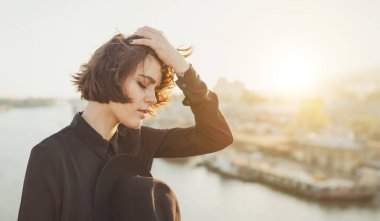 Düşünceli kız aşağı bakıyor, şapkasını elinde tutuyor, saçlarına dokunuyor. Şehir panorama arka planı. Arka ışık. Boş kopya alanı olan uzun yatay afiş. Duygusal açıdan güzel bir portre 