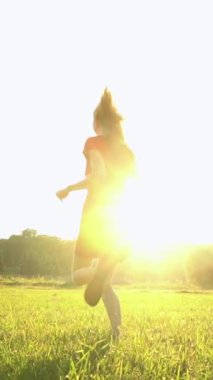 Kısa kırmızı elbiseli sarışın kadın yeşil yaz çayırında mutlu bir şekilde koşan küçük köpeği takip etti. Parlak arka plan güneş ışınları kameraya yansıyor. Yaz hatıraları atmosferi. Yavaş çekim dikey video görüntüleri