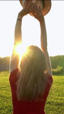 Sarışın, kırmızı elbiseli, hasır şapkalı, elleri havada, parlak güneşe doğru bakan bir kadın. Mutlu bir gülümseme Parktaki doğada güneşli bir yaz. dikey video görüntüleri