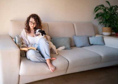 Siyah beyaz köpeğiyle kanepeye oturmuş kitap tutan mutlu kıvırcık kadın. Kot pantolon. Evdeki evcil hayvan arkadaşınla gevşeme zamanı. Güneşli gündüz odası