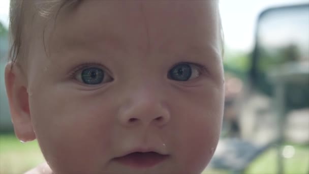 一个可爱的高加索女孩的特写 大漂亮的婴儿眼睛 一个可爱的婴儿的肖像 看着相机 坐在他母亲的怀里 背靠着一片绿色的田野 — 图库视频影像